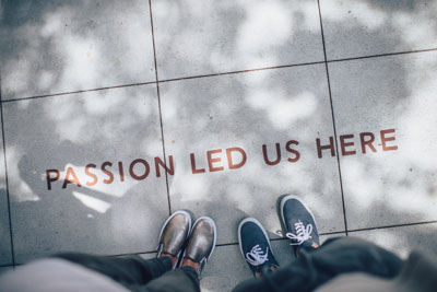 Two pairs of shoes standing on pavement with text: 'PASSION LED US HERE'.